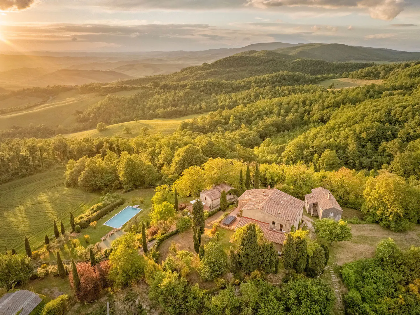 Aerial view of a Tuscan villa with a pool, surrounded by green hills and trees at sunset.
