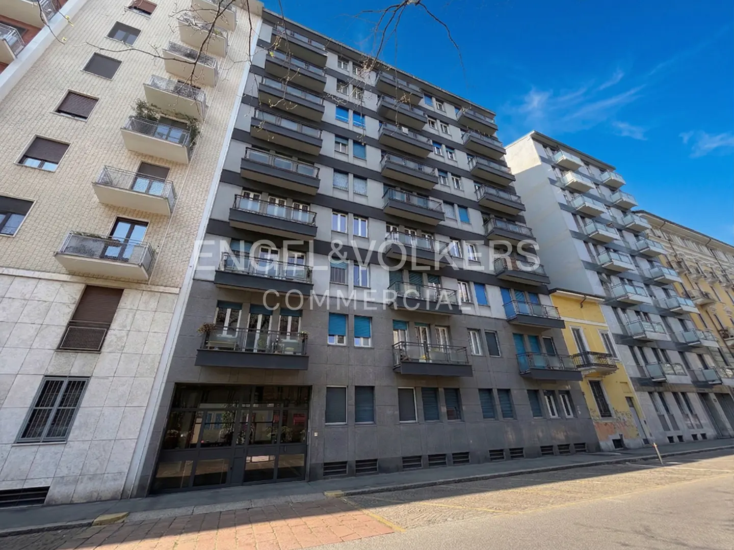 Exterior view of a tall, gray apartment building with balconies under a blue sky. Engel & Volkers Commercial logo is superimposed on the building.