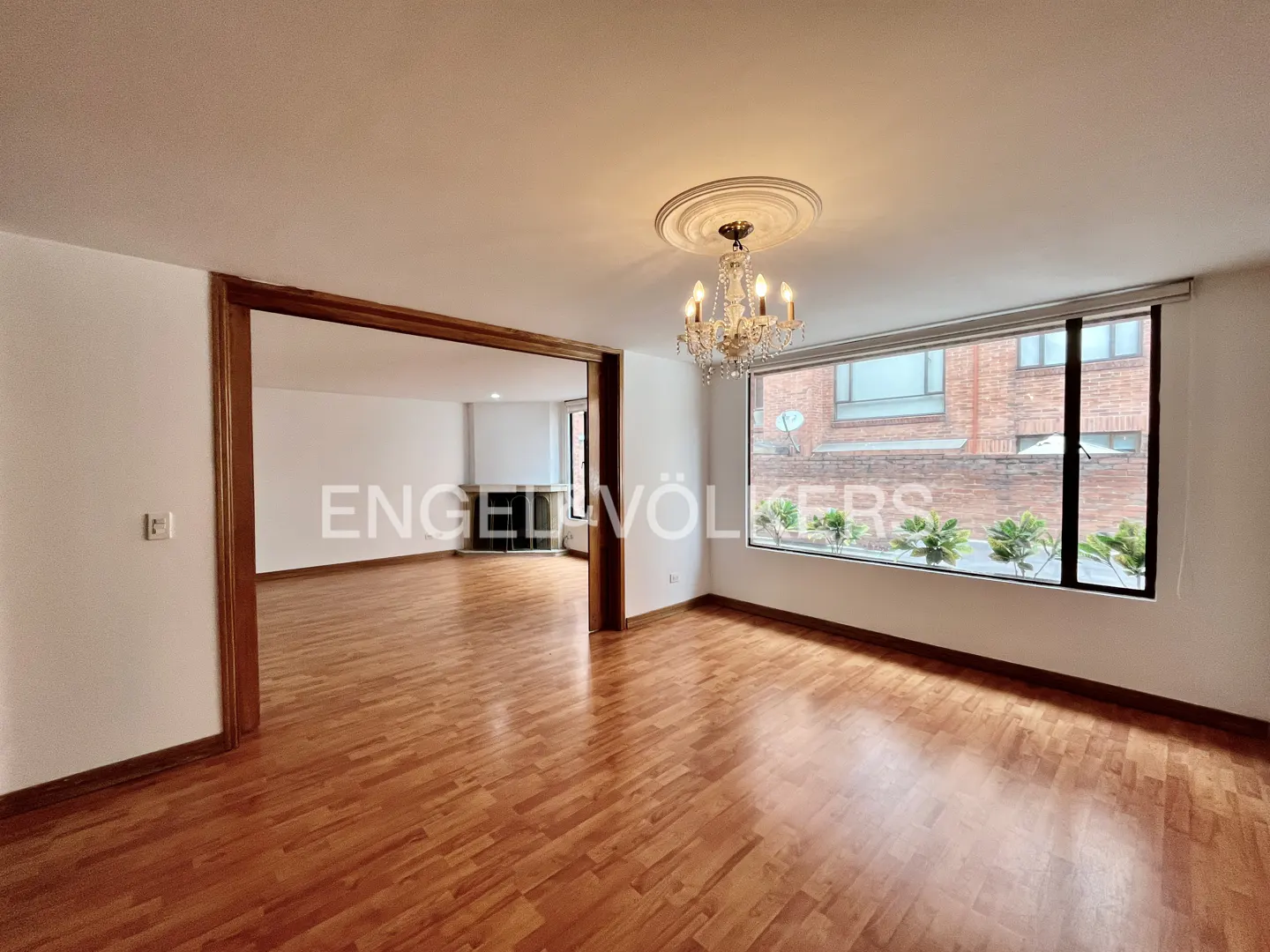 Bright, empty living room with hardwood floors, white walls, and a chandelier. A large window overlooks a brick building and greenery.