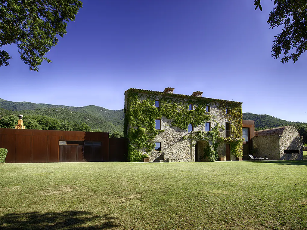 Exterior of a stone house covered in ivy, with a green lawn, blue sky, and mountains in the background.