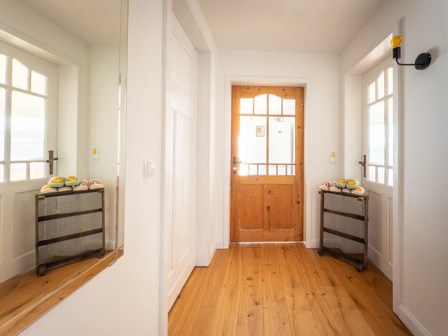 Bright hallway with wood floors, white walls, and a wooden front door. A mirror reflects a white door and a shoe rack with slippers.