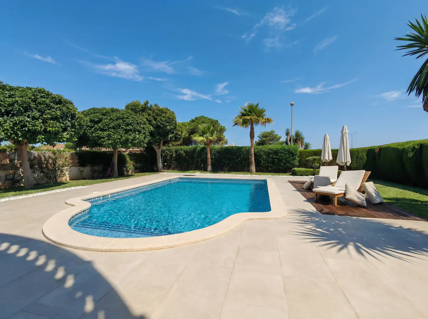 A kidney-shaped pool with blue water, surrounded by a beige patio, green grass, trees, and two lounge chairs.