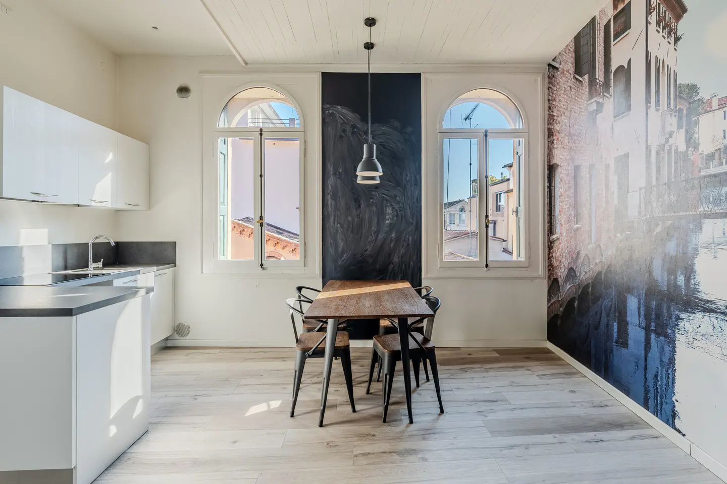 Bright kitchen with white cabinets, wood table and chairs, and a Venice canal mural. Arched windows let in natural light.