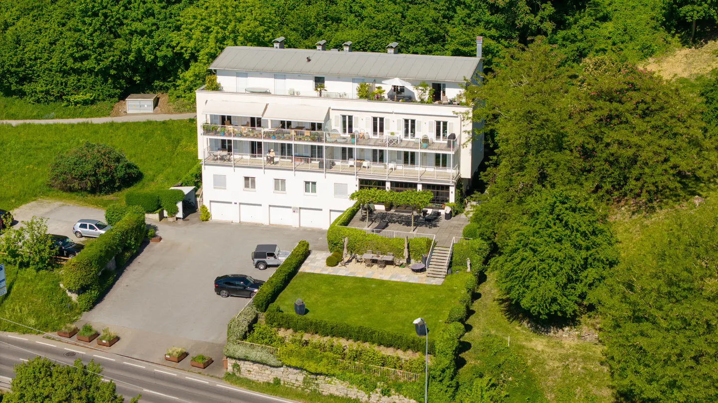 Aerial view of a white multi-story apartment building with balconies, a green lawn, and a parking area surrounded by trees.