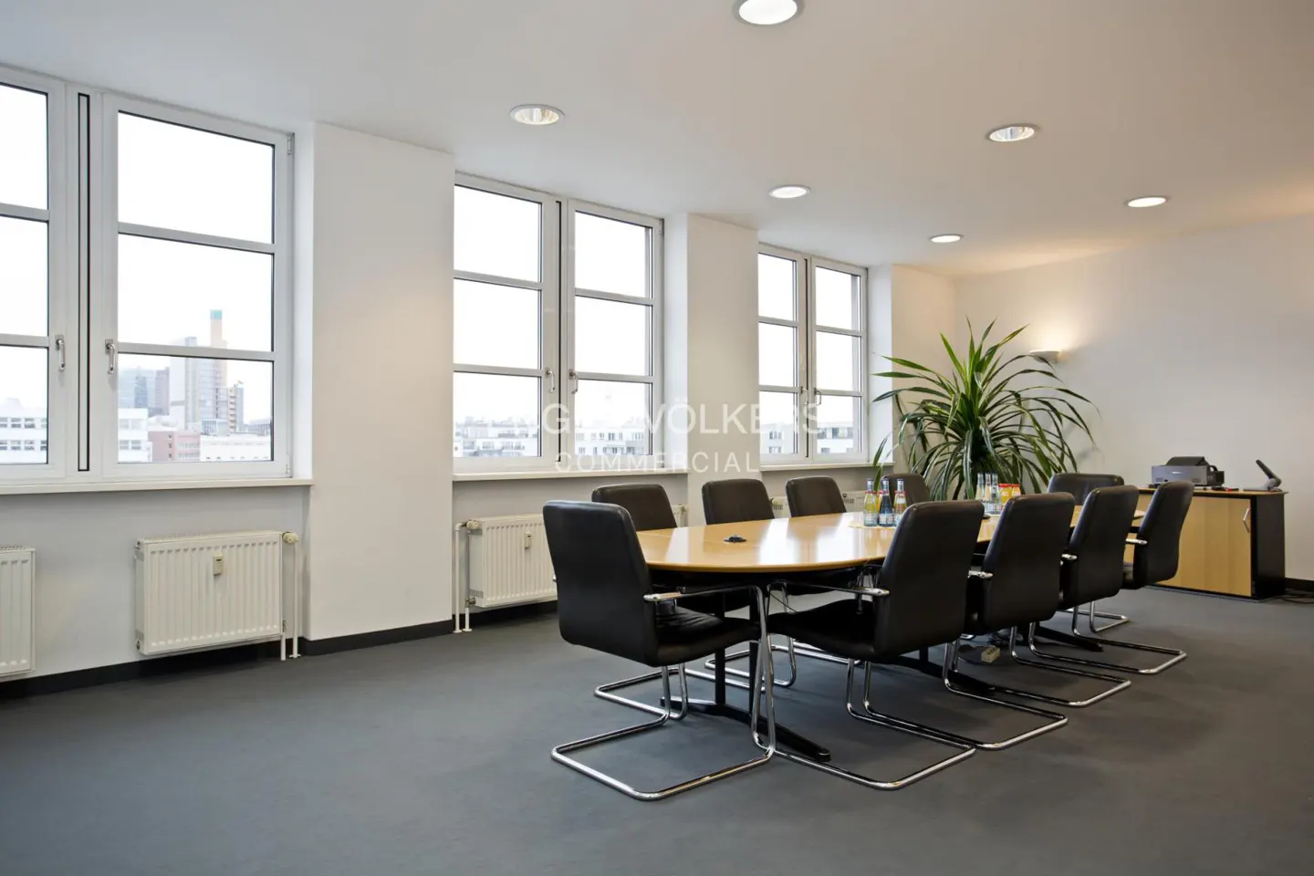 Bright conference room with a long oval table, black chairs, and large windows overlooking a city. A plant and desk are in the background.
