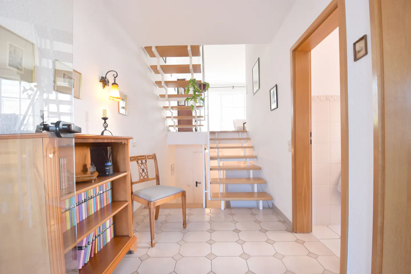 Bright foyer with white walls, tiled floor, and wooden accents. A bookcase, chair, and stairs are visible. A door leads to a white-tiled bathroom.