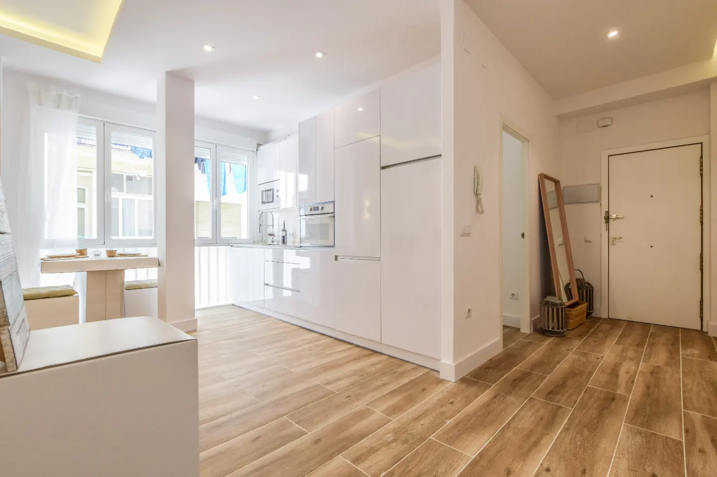 Modern white kitchen with wood-look tile floors, a small dining area, and an entryway with a floor mirror and white door.