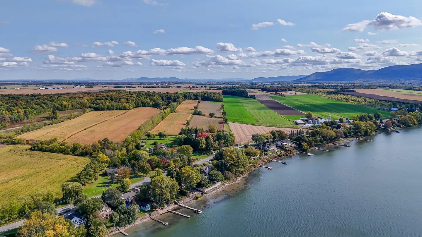 Aerial view of waterfront homes, docks, and farmland under a blue sky with scattered clouds. Mountains are visible in the distance.