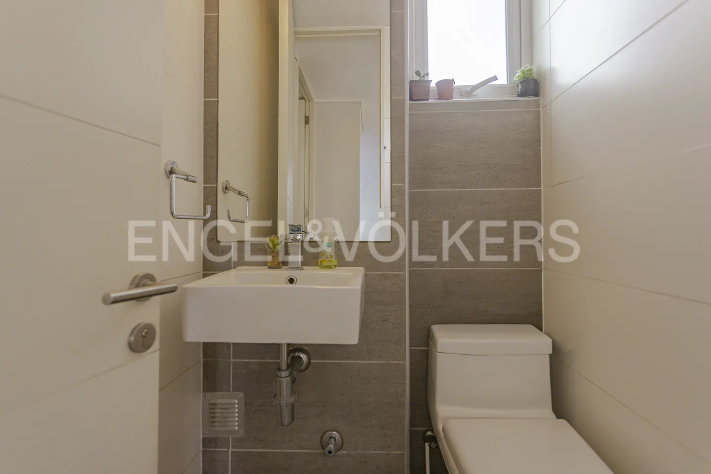 A small bathroom with a white toilet, sink, and door. Gray and white tiles line the walls. A mirror hangs above the sink.