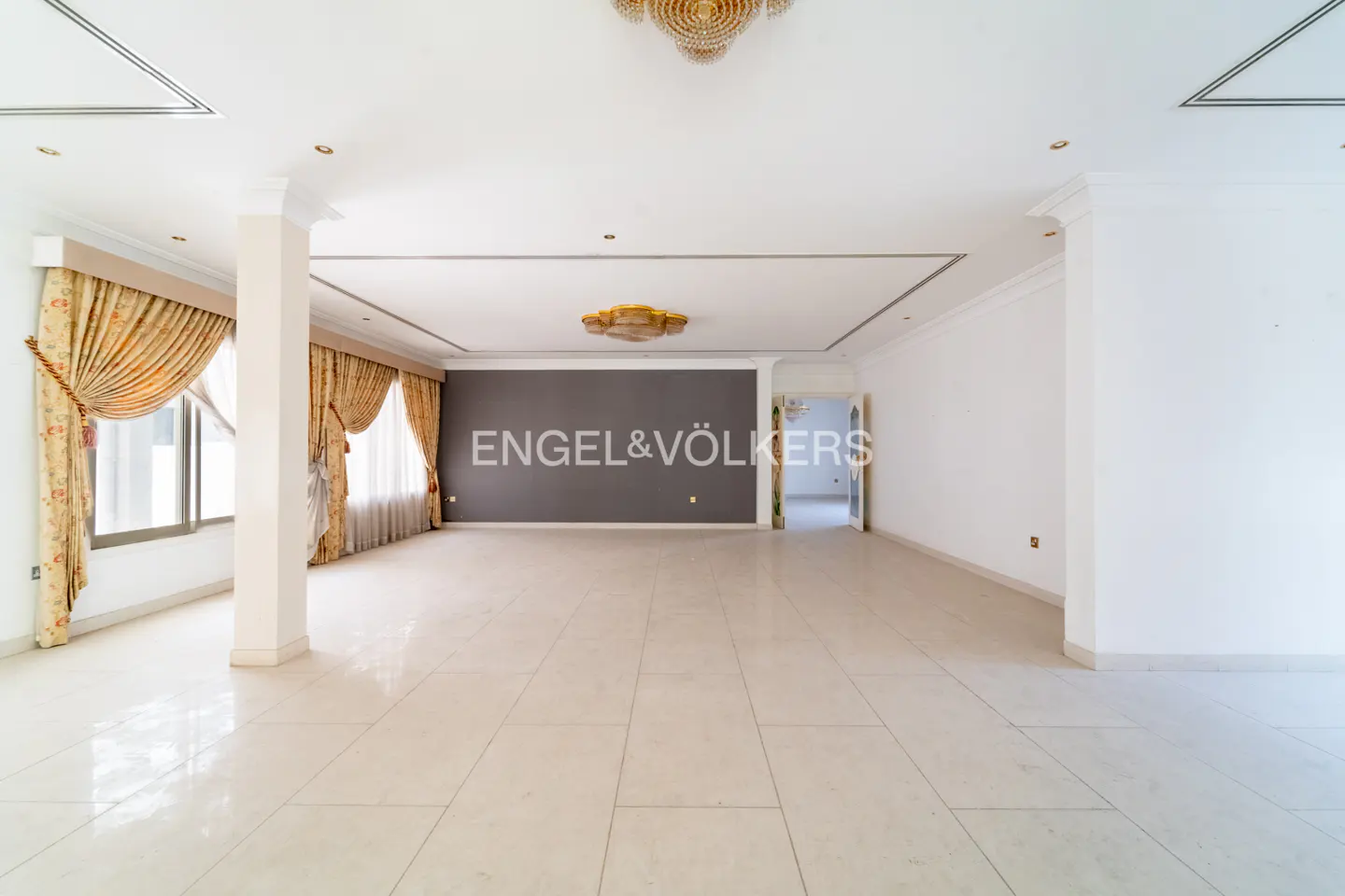 A spacious, empty room with beige tile flooring, white walls, and gold curtains. The Engel & Völkers logo is on a gray wall.