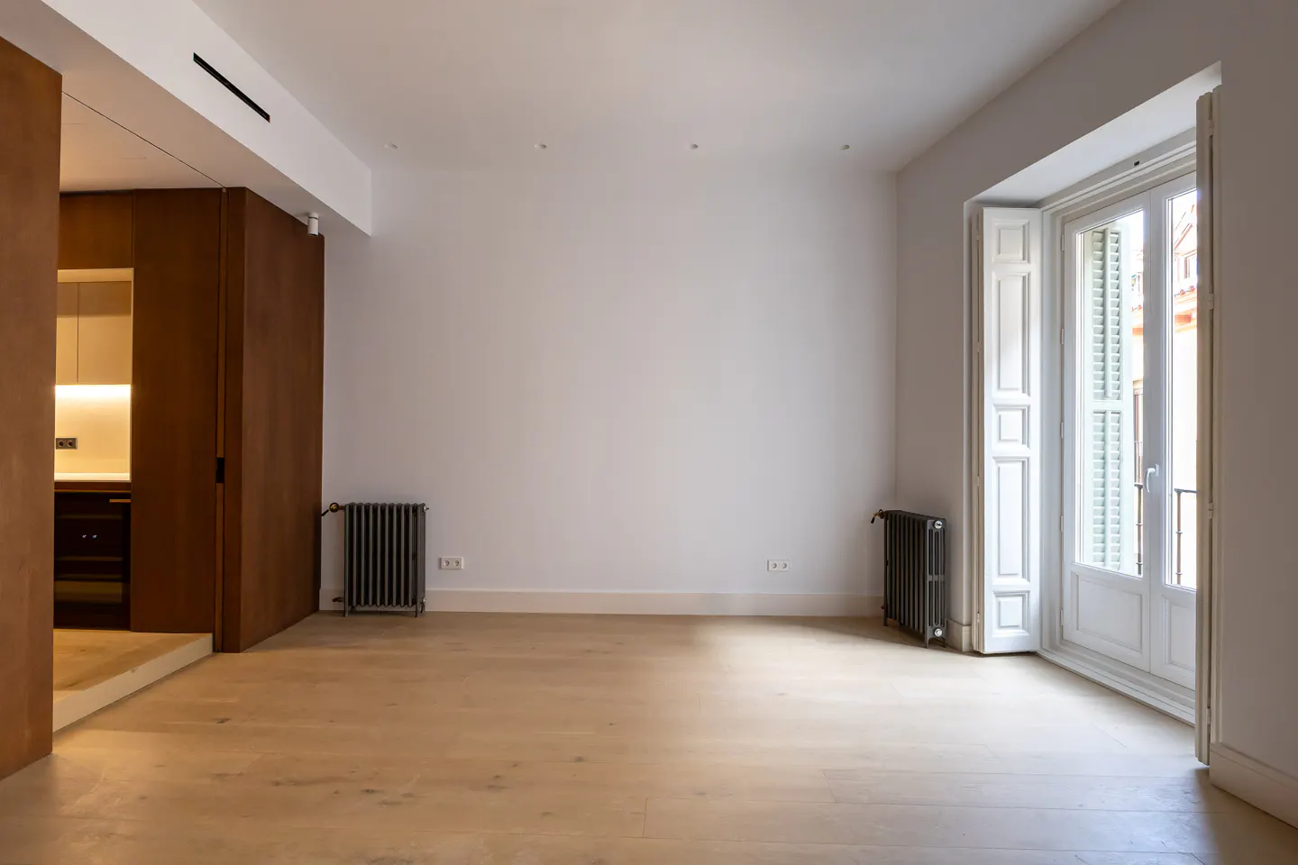 Bright, empty room with wood floors, white walls, and a white shuttered door to a balcony. A kitchen is visible through a doorway.