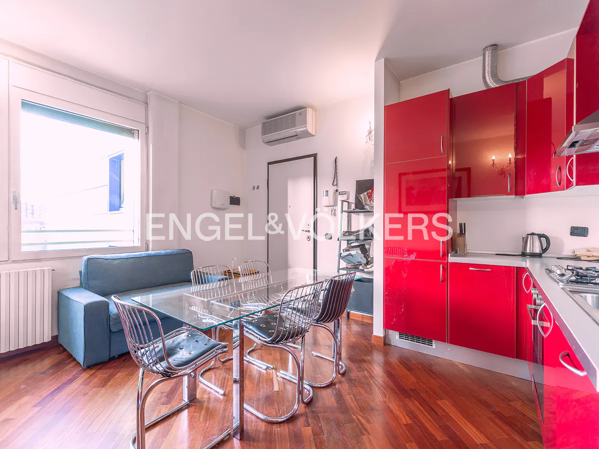A modern apartment interior with a glass table, metal chairs, a blue sofa, and red kitchen cabinets.