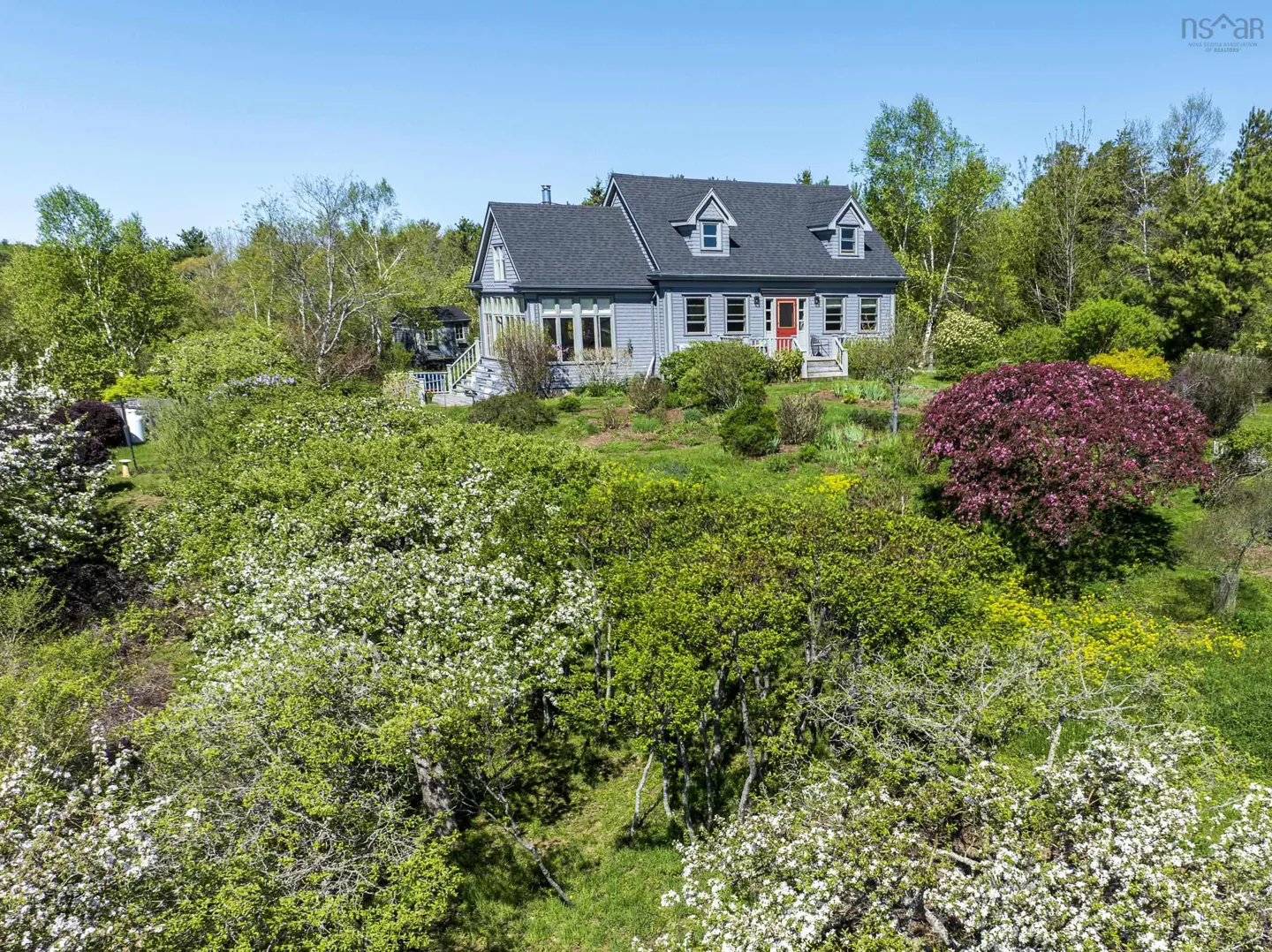 Light blue house with a grey roof and red door, surrounded by lush green trees and flowering bushes.
