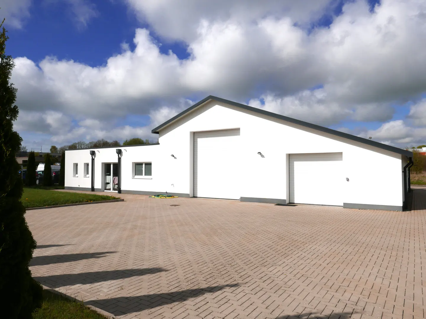 Modern white commercial building with two garage doors and a paved driveway under a bright, cloudy blue sky.