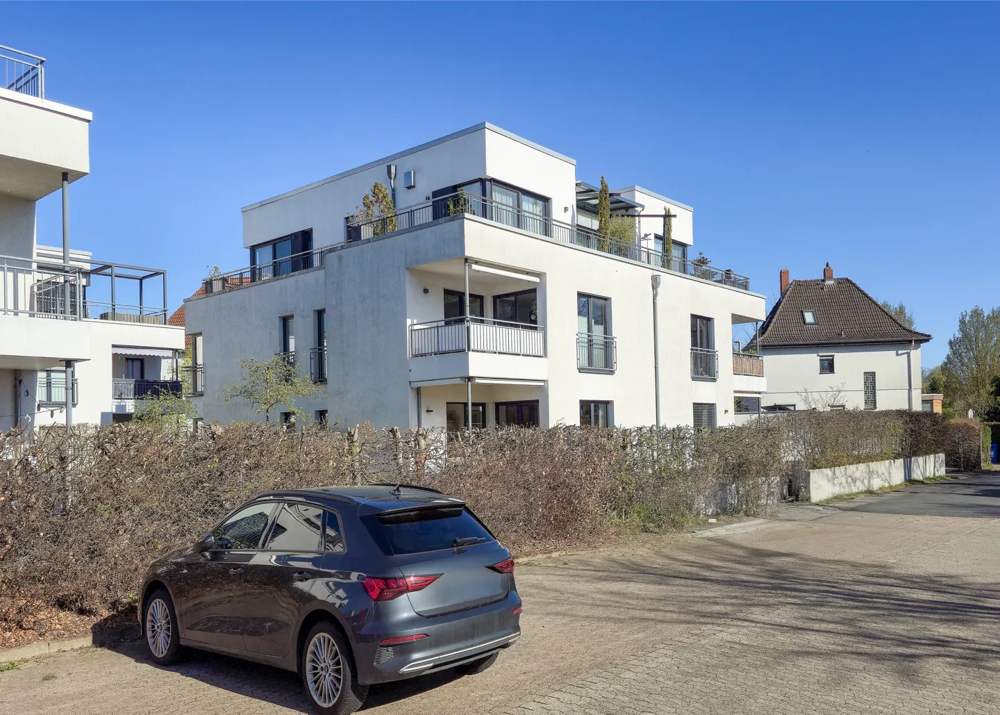 A modern white apartment building with balconies, a gray car parked in front, and a blue sky.