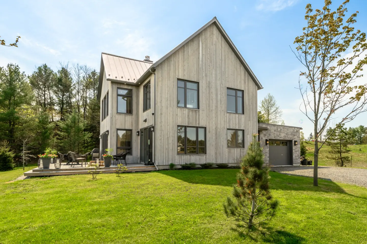 Exterior view of a modern farmhouse with gray wood siding, black framed windows, and a metal roof, surrounded by a green lawn and trees.
