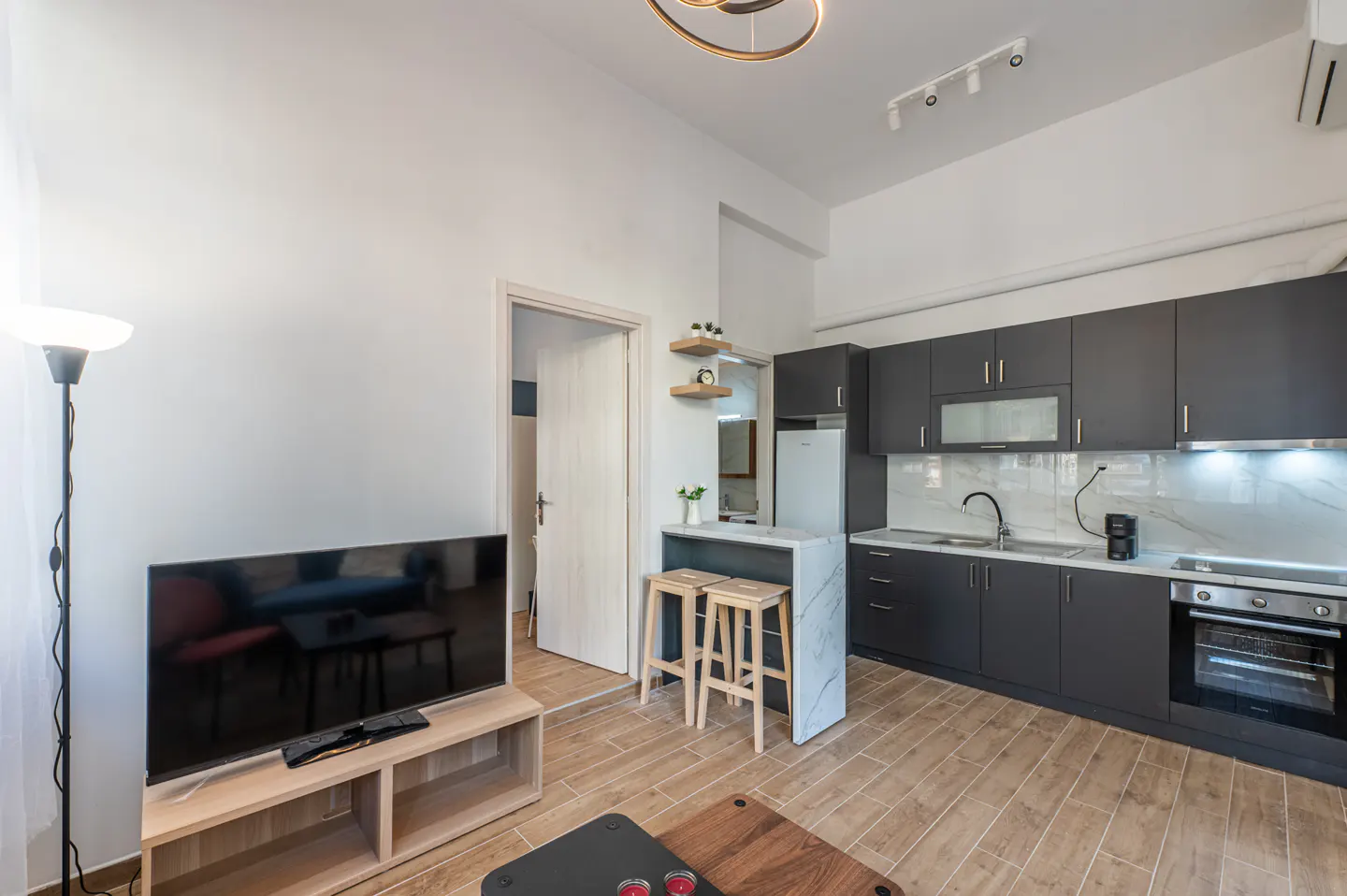 A modern apartment interior with a TV, kitchen with gray cabinets, and a breakfast bar with two stools.
