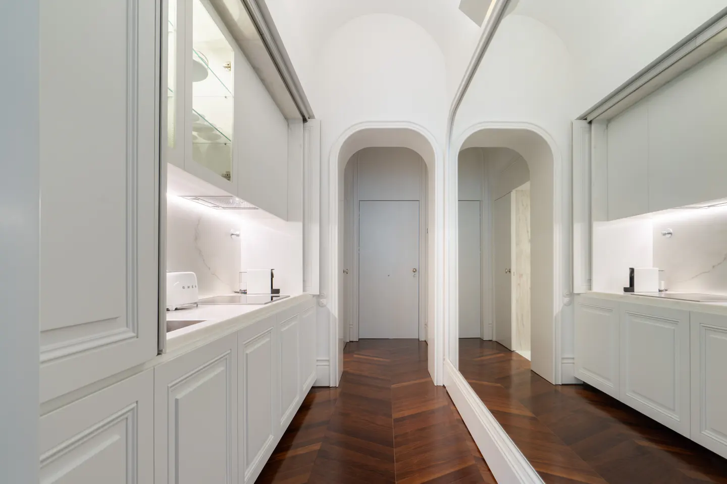Hallway with white cabinets, marble countertops, and a herringbone wood floor. A mirror reflects the hallway.