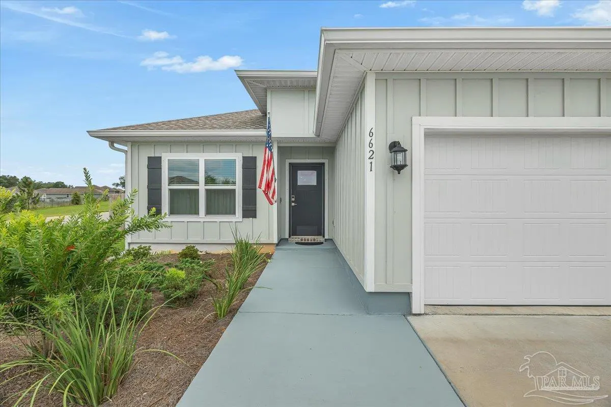 Light gray house exterior with a white garage door, dark front door, American flag, and a concrete walkway.