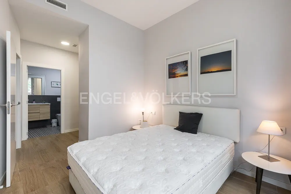 Bedroom with white walls, wood floors, and a white bed with a black pillow. Two framed pictures hang above the bed. Bathroom visible through doorway.