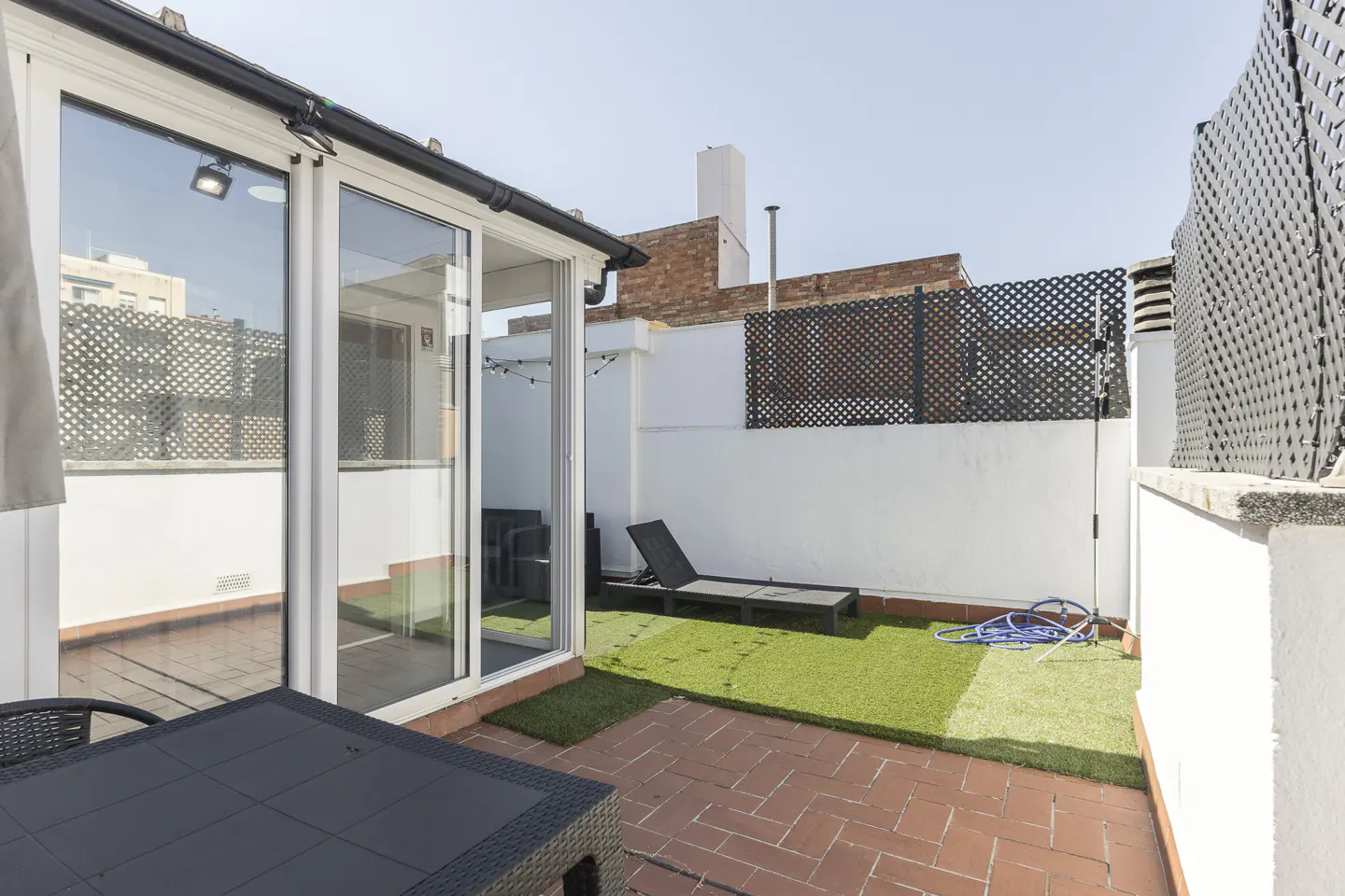 A rooftop patio with a black table, lounge chair, and artificial grass. White walls and a black lattice fence surround the space.