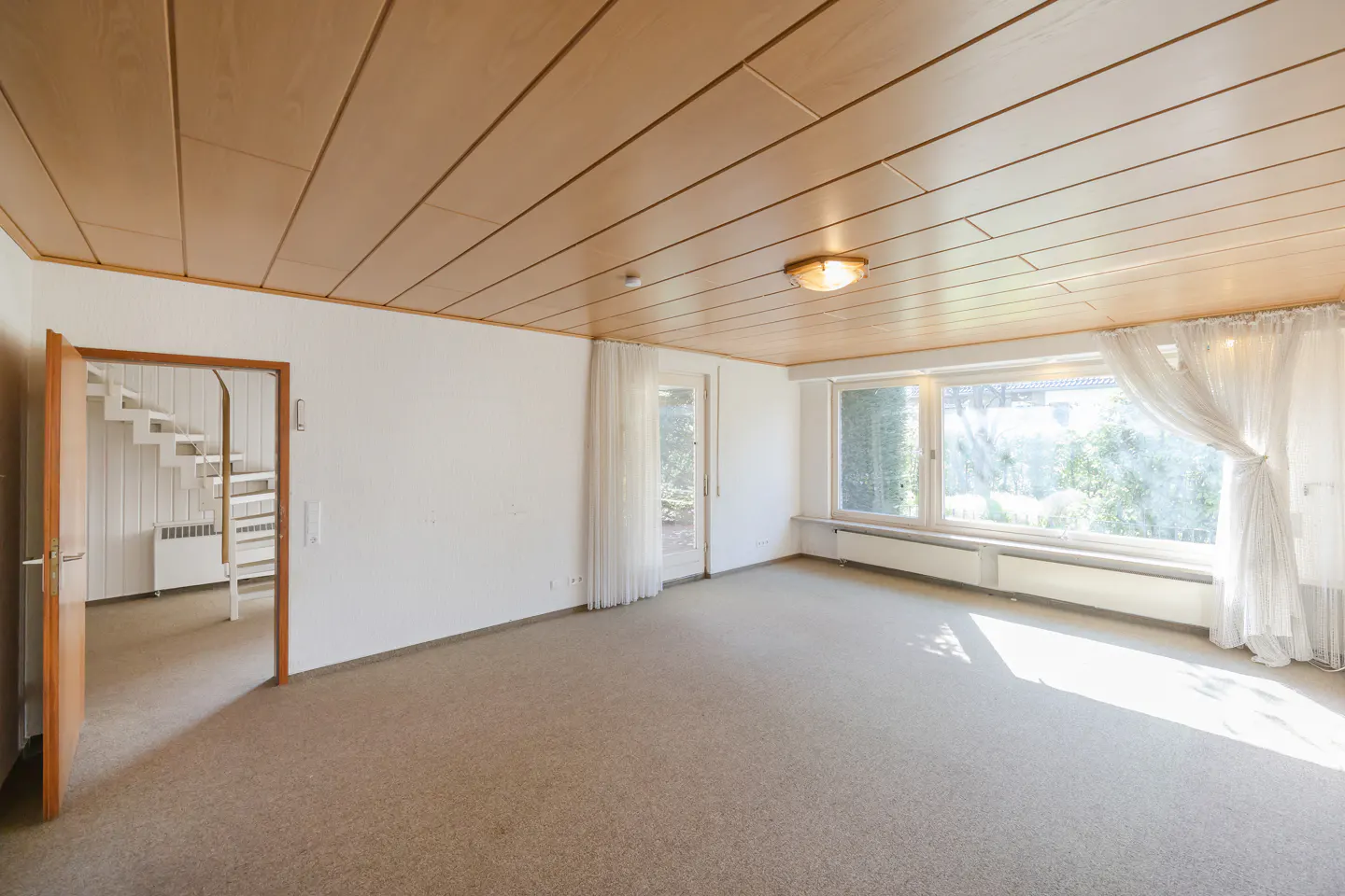 Bright, empty room with beige carpet, wood ceiling, and large windows with sheer white curtains. A white staircase is visible through an open door.