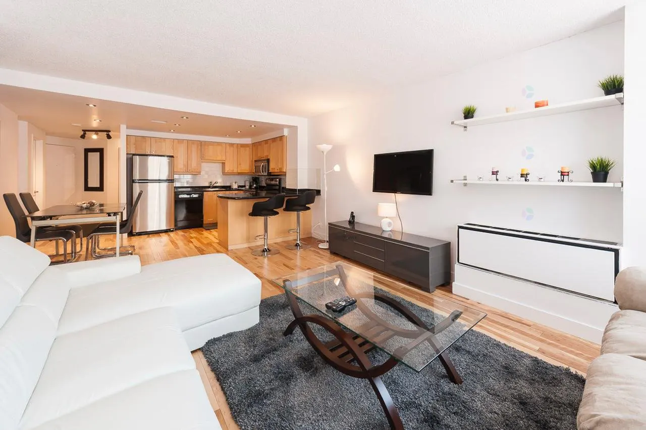 A modern apartment living room with a white sofa, glass table, and open kitchen. Shelves with plants and decor add a touch of nature.