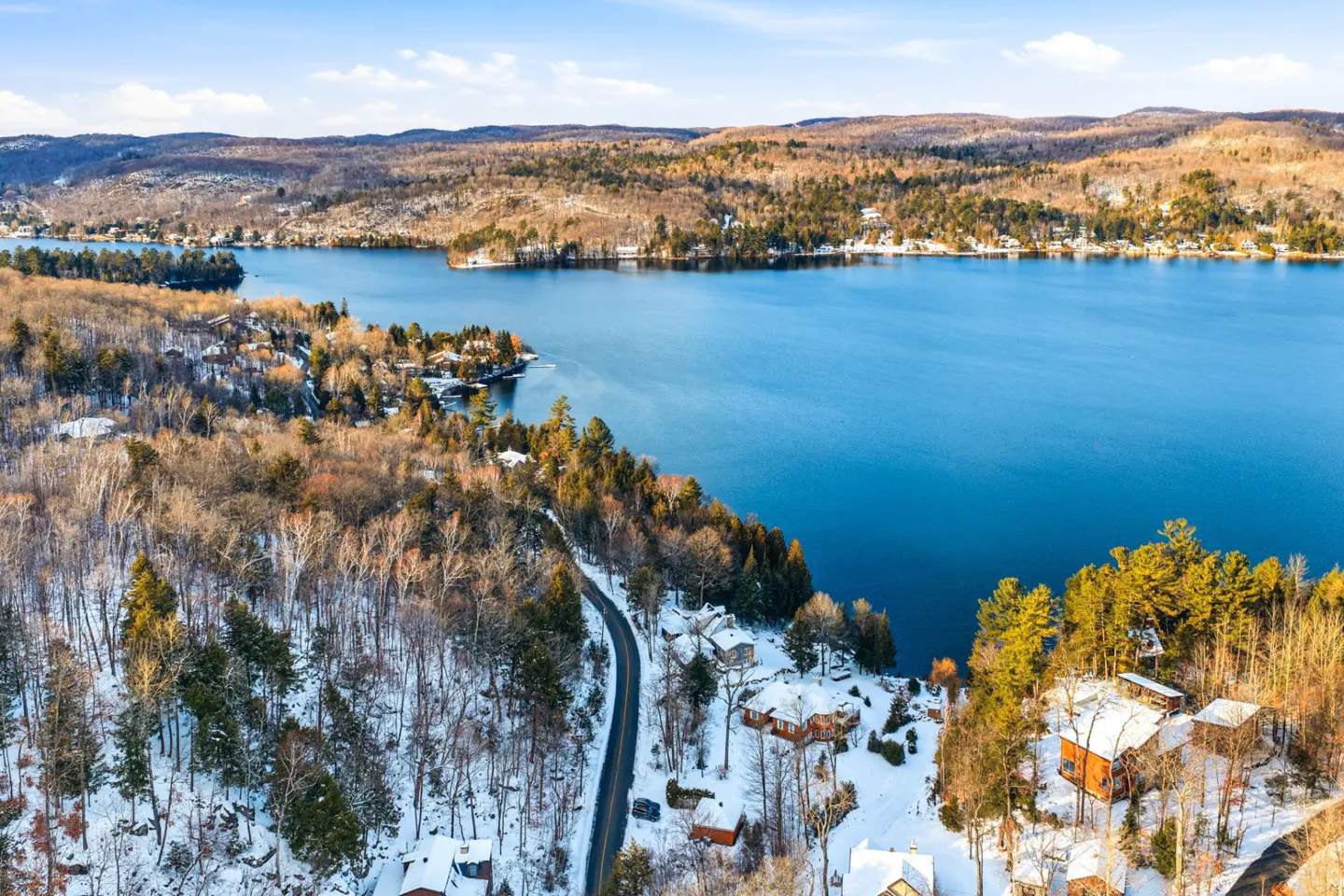 Aerial view of a blue lake surrounded by snow-dusted hills and trees, with houses nestled along the shoreline and a road winding through the landscape.