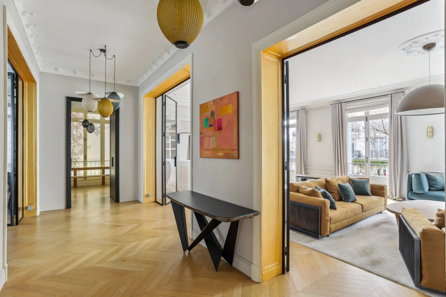 Interior view of a modern apartment with herringbone wood floors, gray walls, and gold trim. A black console table sits beneath an abstract painting. Sofas are visible in the living room.