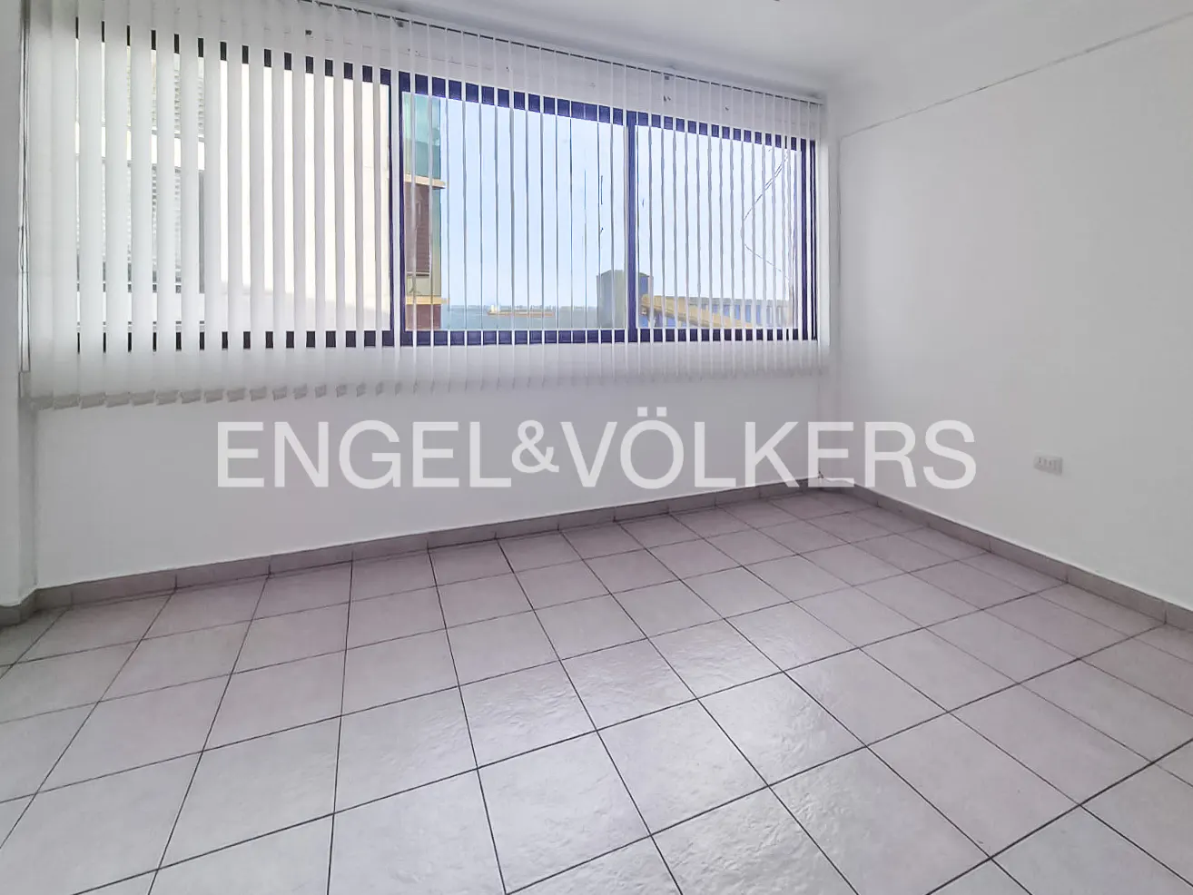 Empty room with white walls, tiled floor, and large window with vertical blinds. Engel & Völkers logo on the wall.