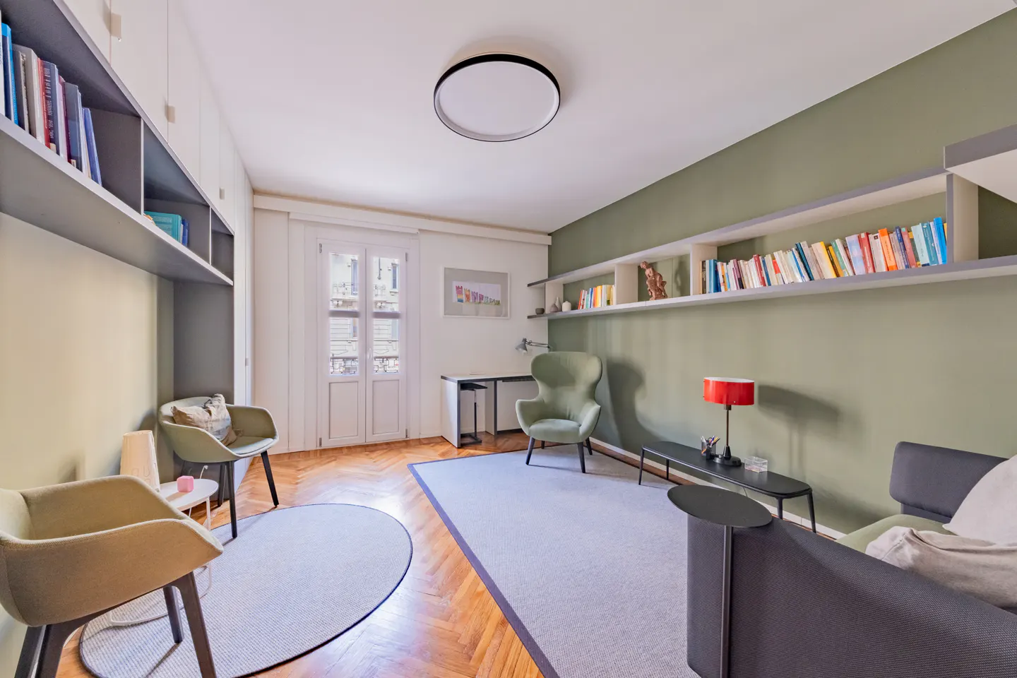A living room with parquet floors, a gray rug, a gray sofa, and green walls with bookshelves.