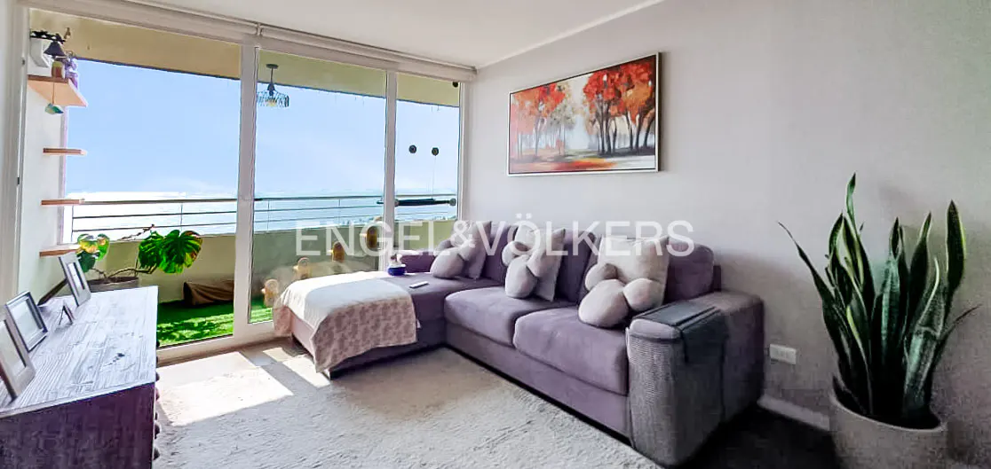 Living room with a lilac sofa, white rug, and balcony overlooking the ocean. A painting hangs on the wall.