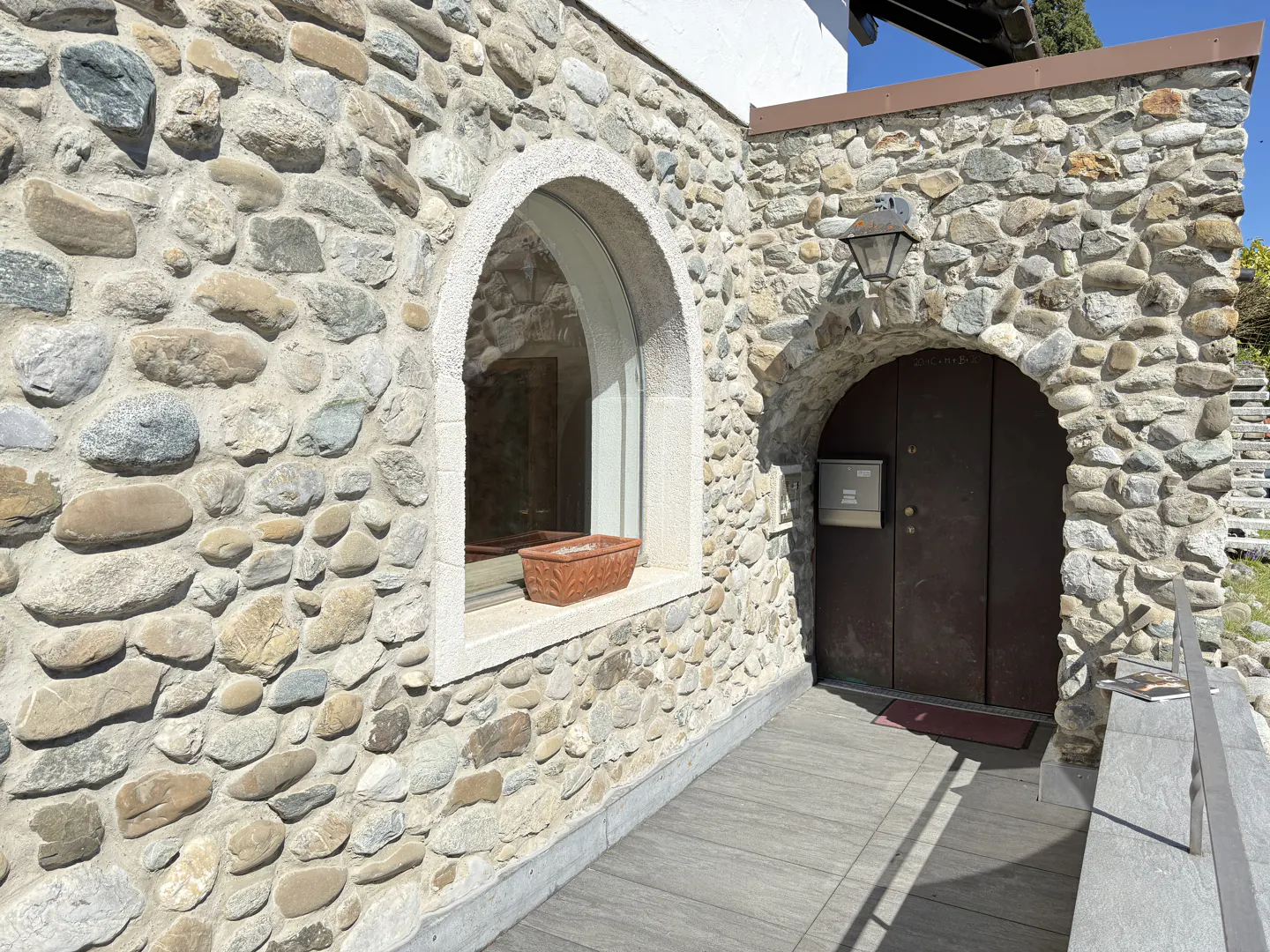 Stone house exterior with arched window and door. A brown planter sits on the window sill. A lantern hangs above the door.
