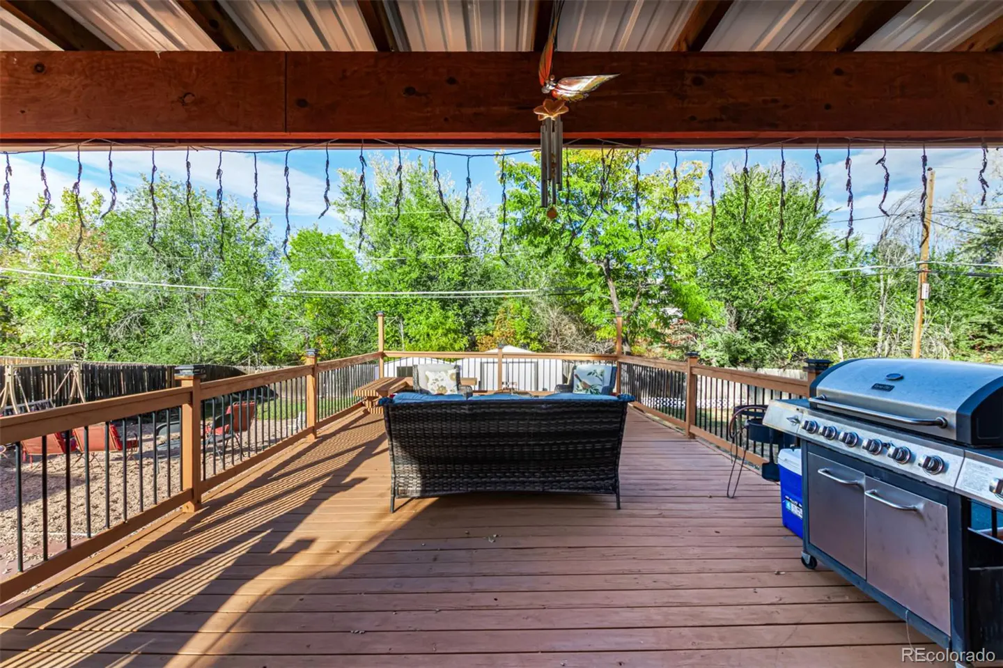 Covered wooden deck with a wicker sofa, grill, and green trees in the background.