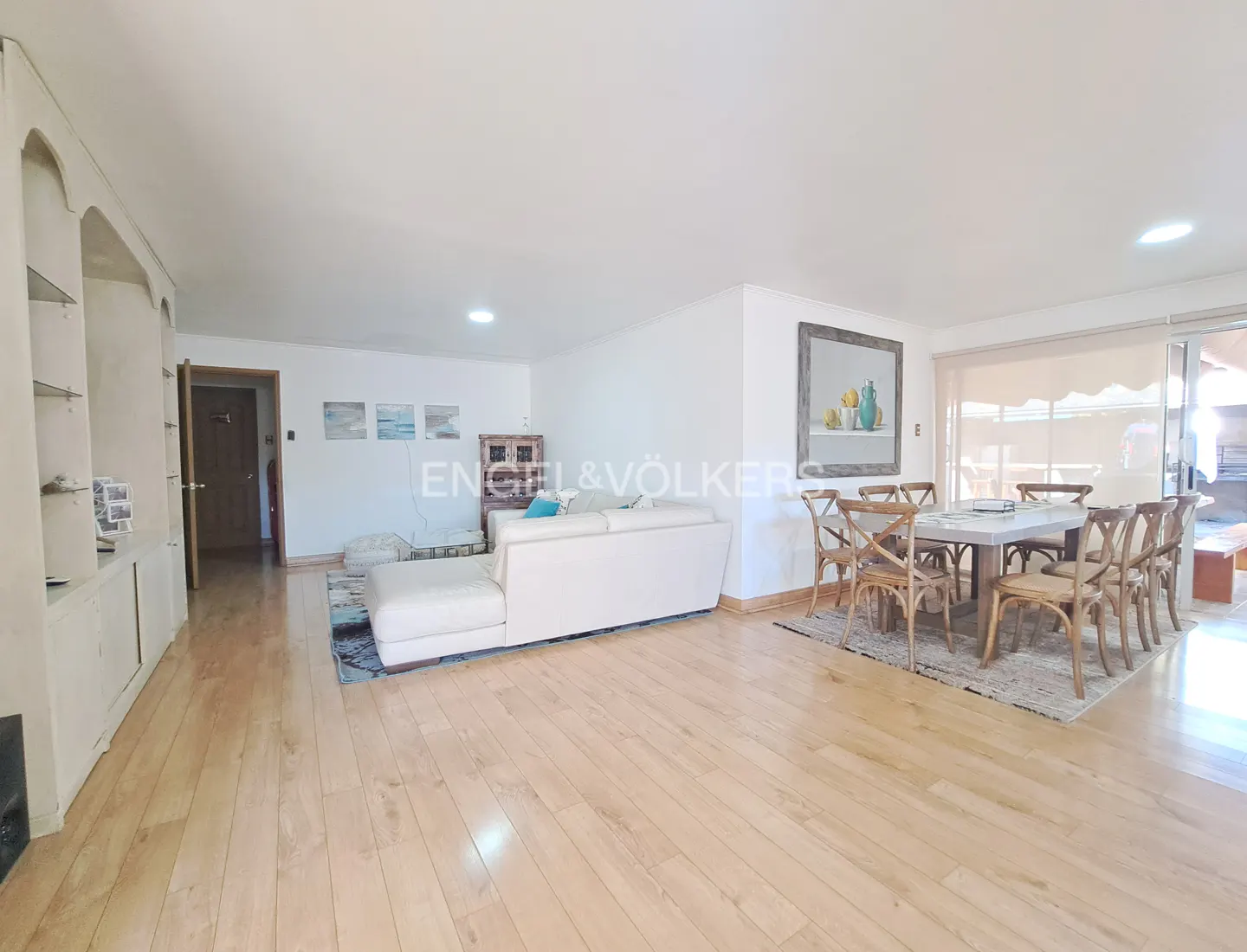Bright, open-plan living and dining area with hardwood floors. White sectional sofa, dining table with eight chairs, and built-in shelving.