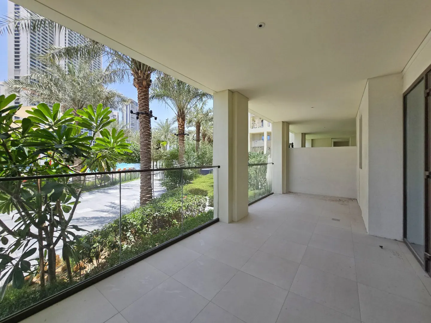 A bright, empty balcony with white tile flooring and a glass railing overlooks a lush green garden with palm trees.