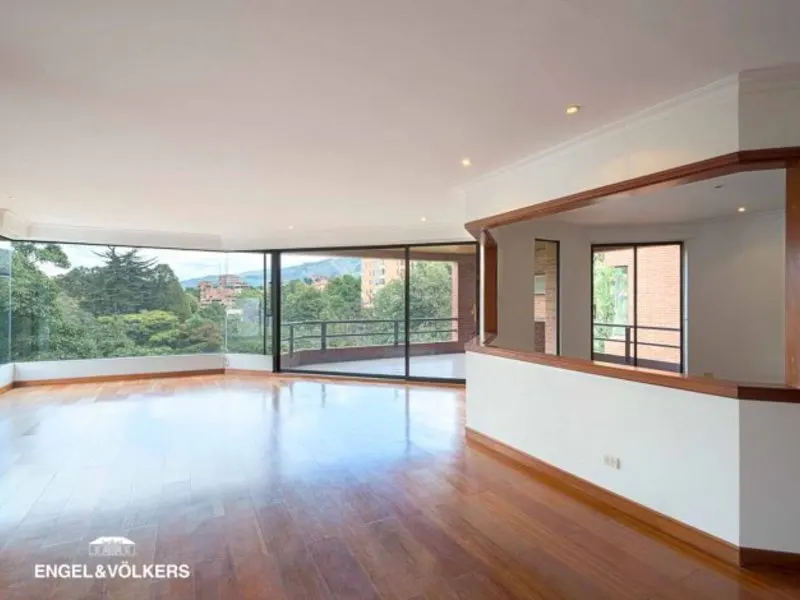 Bright, empty living room with hardwood floors and large windows overlooking trees and mountains. White walls and a partial wall with a wood trim.