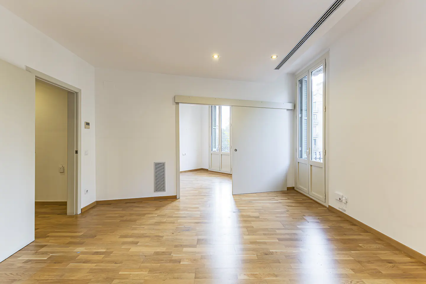 Bright, empty room with light wood floors, white walls, a sliding door, and a doorway leading to another sunlit room.