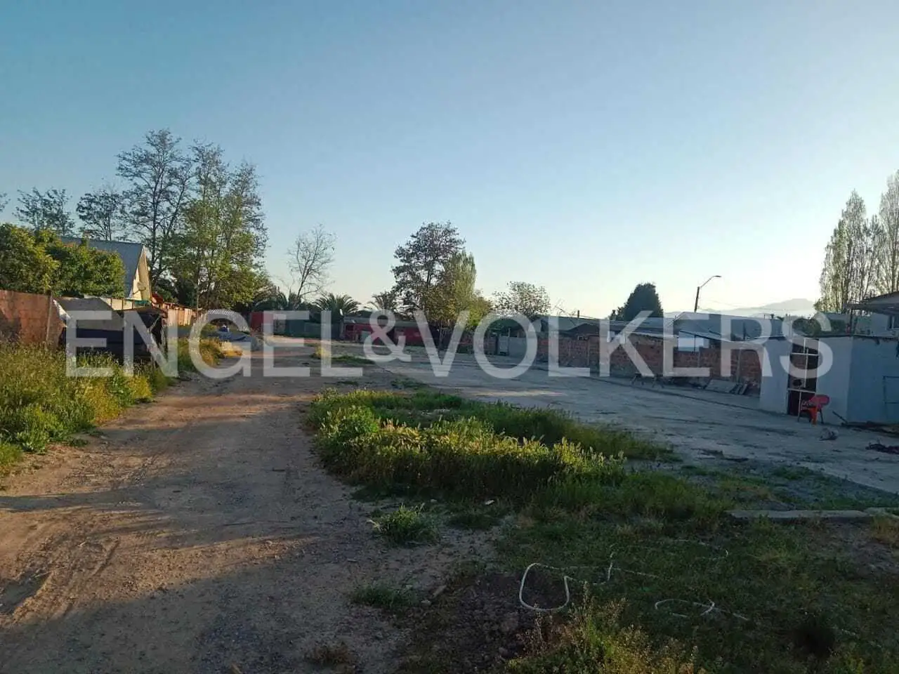 A vacant lot with a dirt road, grass, trees, and buildings under a blue sky. The Engel & Volkers logo is superimposed on the image.