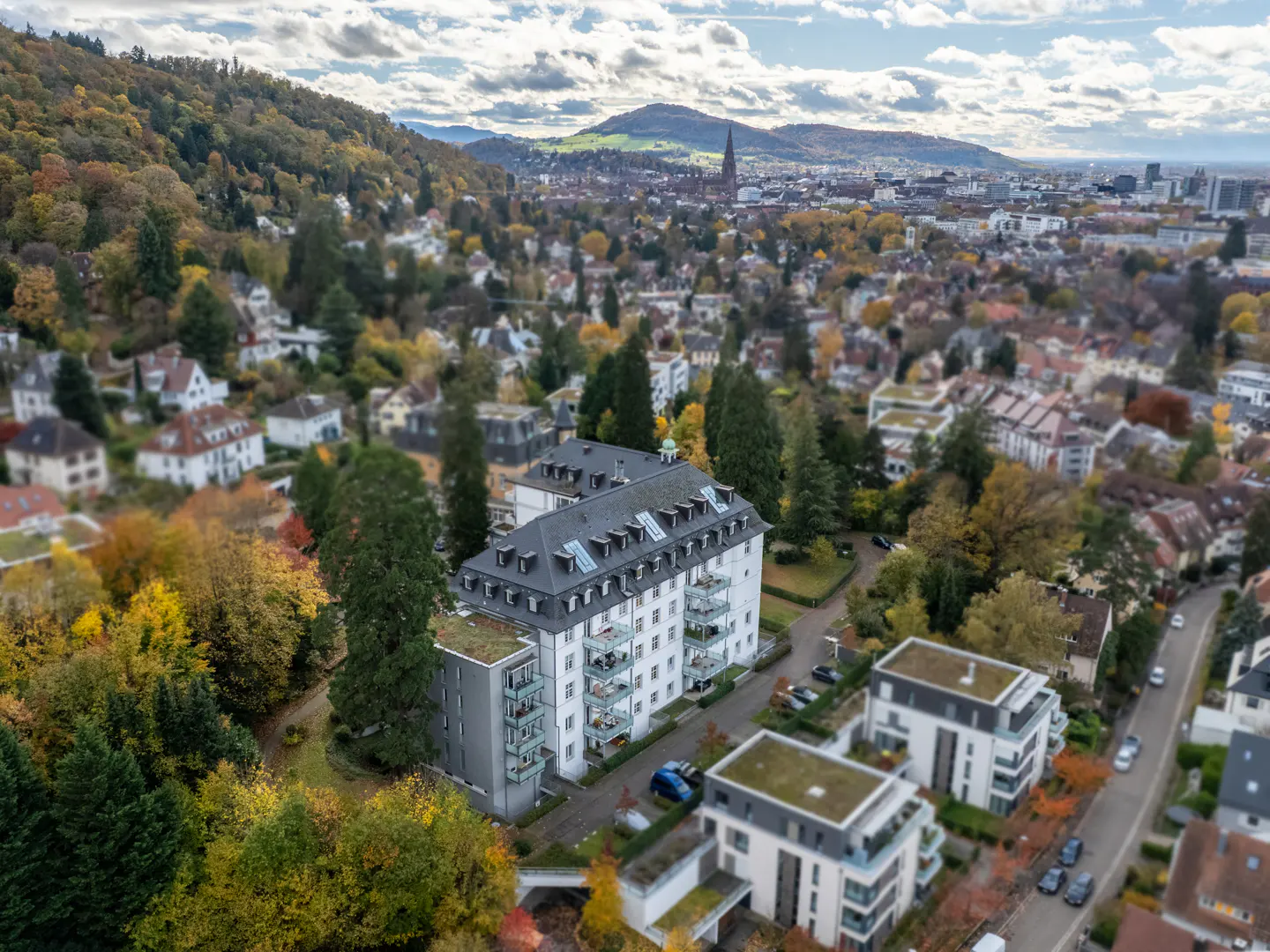 Aerial view of a white apartment building with balconies, surrounded by autumn trees and a cityscape in the background.