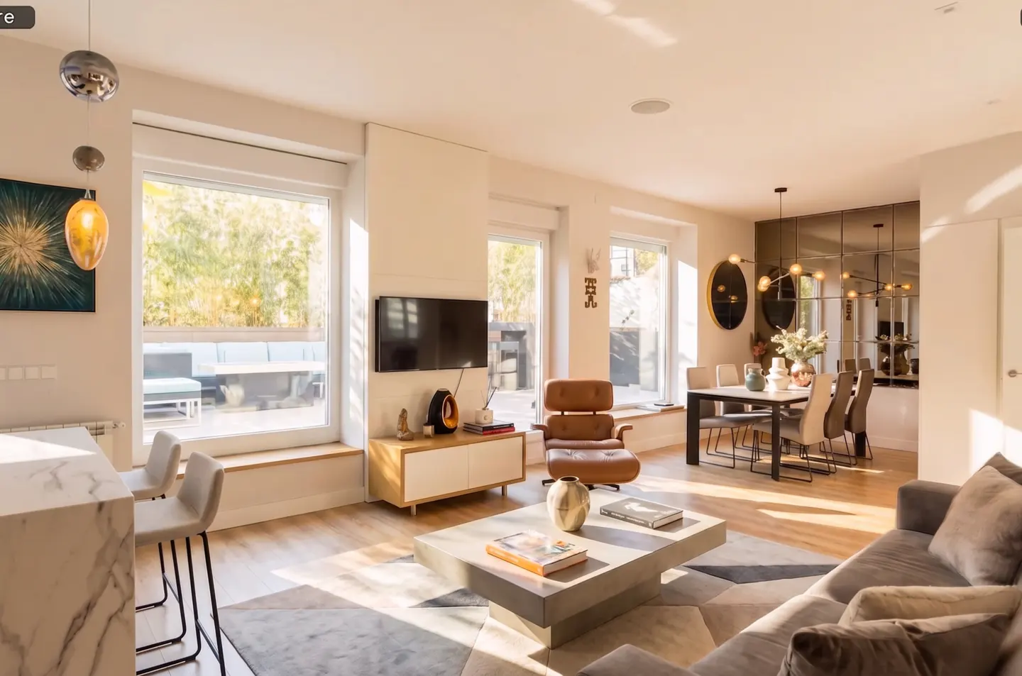 Bright, modern living room with a gray sofa, concrete coffee table, and brown leather chair. Dining area with mirrored wall in the background.