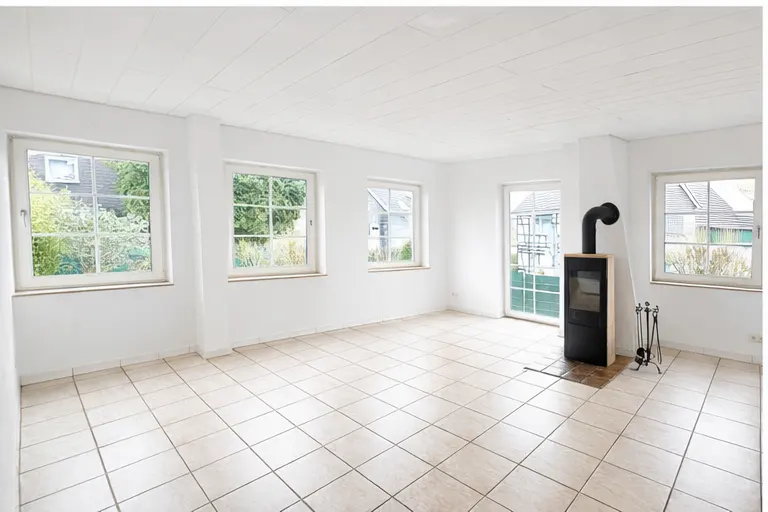 Bright, empty room with white walls, tile floor, and four windows showing green trees. A black wood-burning stove stands near a glass door.