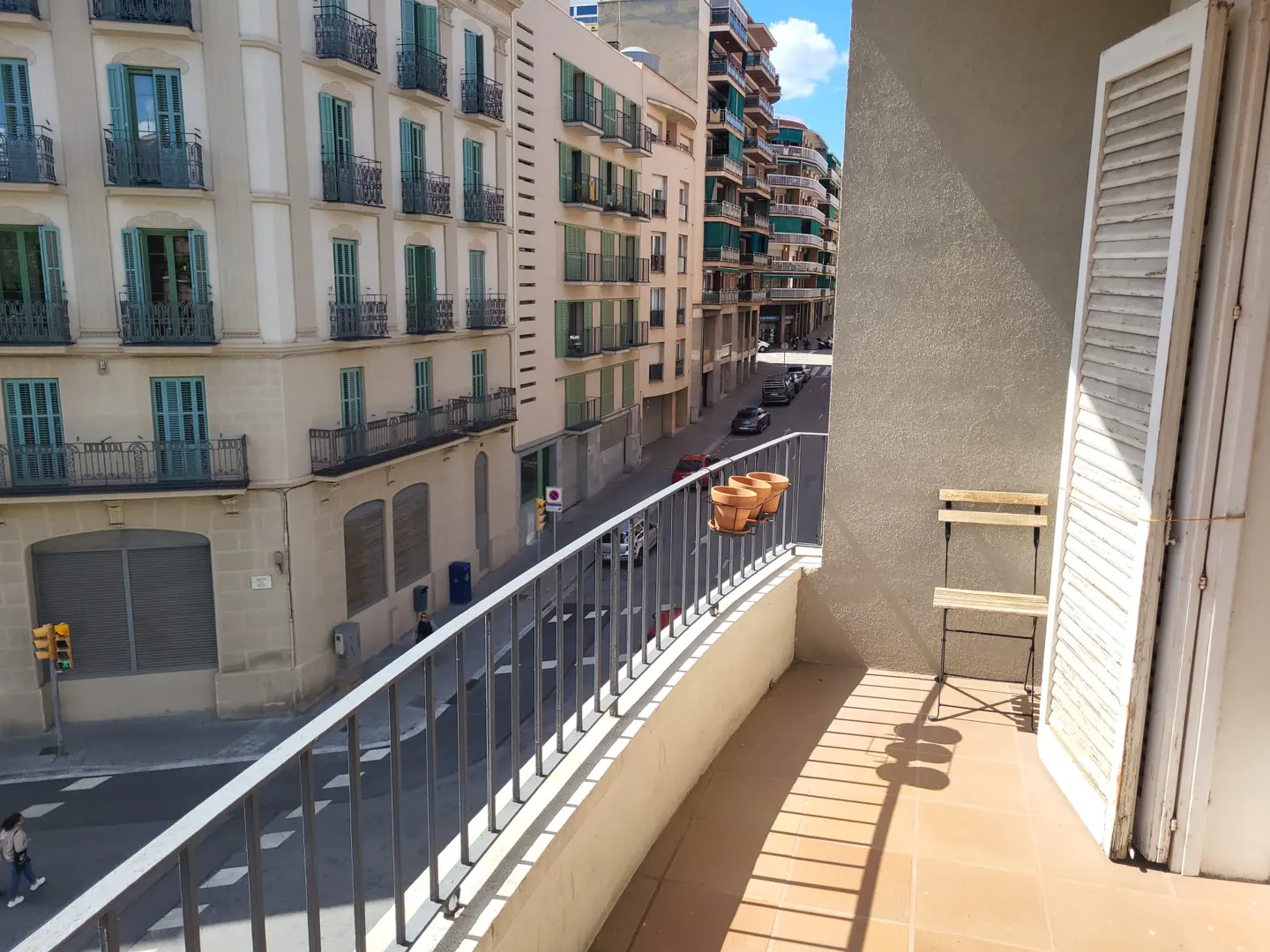 Balcony view of a European city street. A chair and terracotta pots sit on the tiled balcony. White shutters are open.