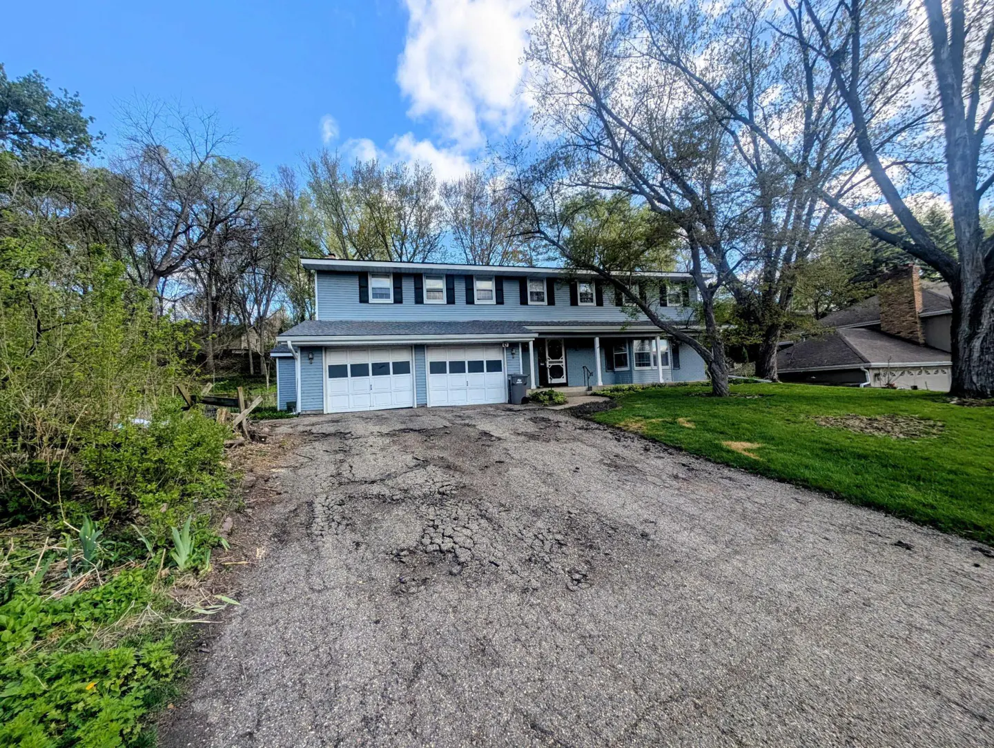 Two-story light blue house with a two-car garage and black shutters. A cracked asphalt driveway leads to the house.