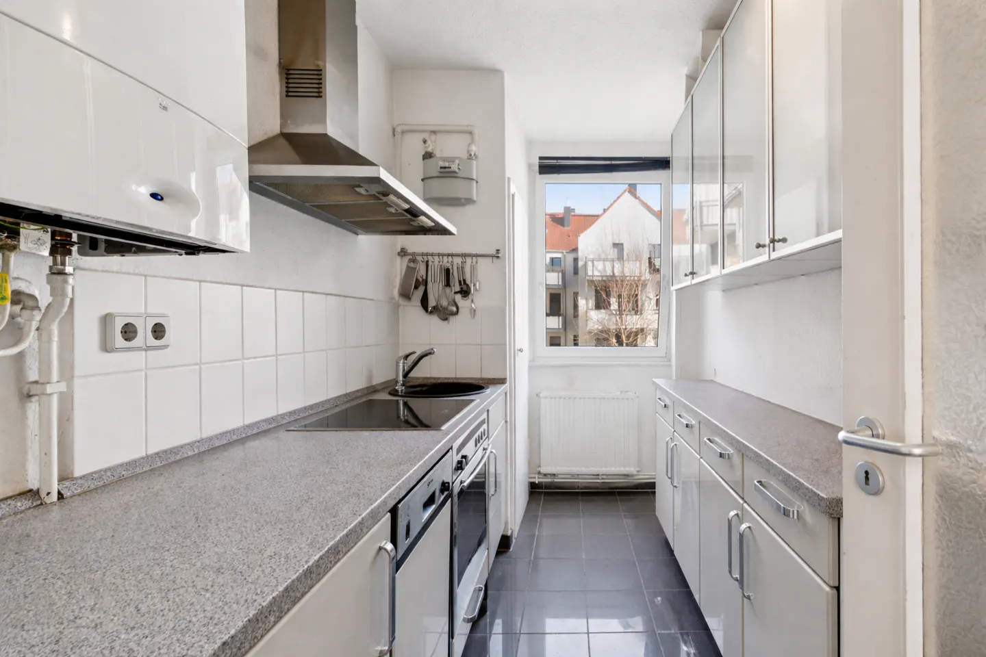 A narrow kitchen with white cabinets, gray countertops, and a stainless steel range hood. A window shows a building exterior.