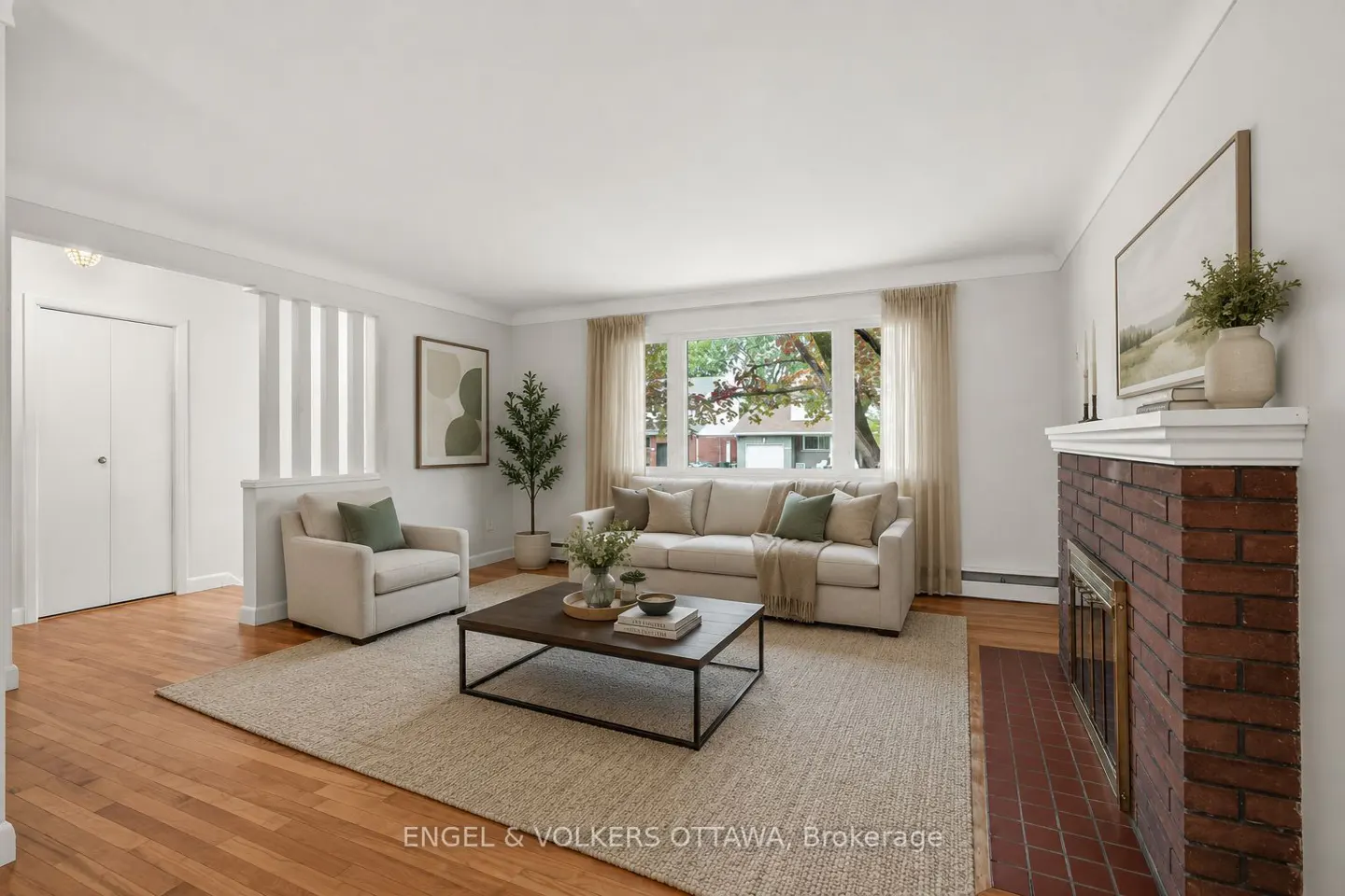 Bright living room with hardwood floors, a beige sofa and armchair, a brick fireplace, and a large window.