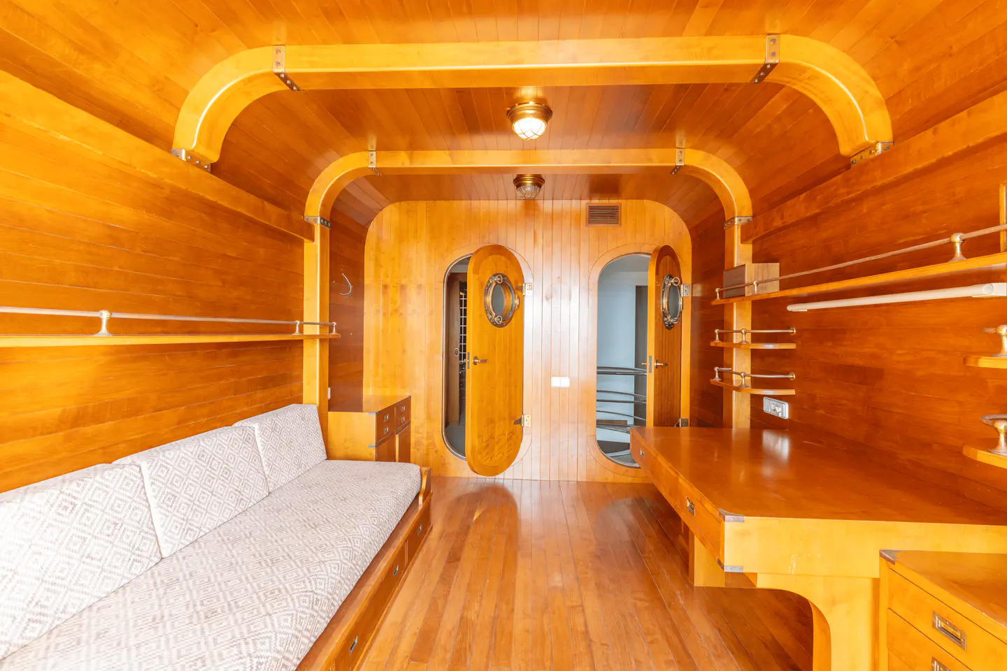 Interior of a boat cabin with warm wood paneling, a patterned sofa, a built-in desk, and two arched doors with portholes.
