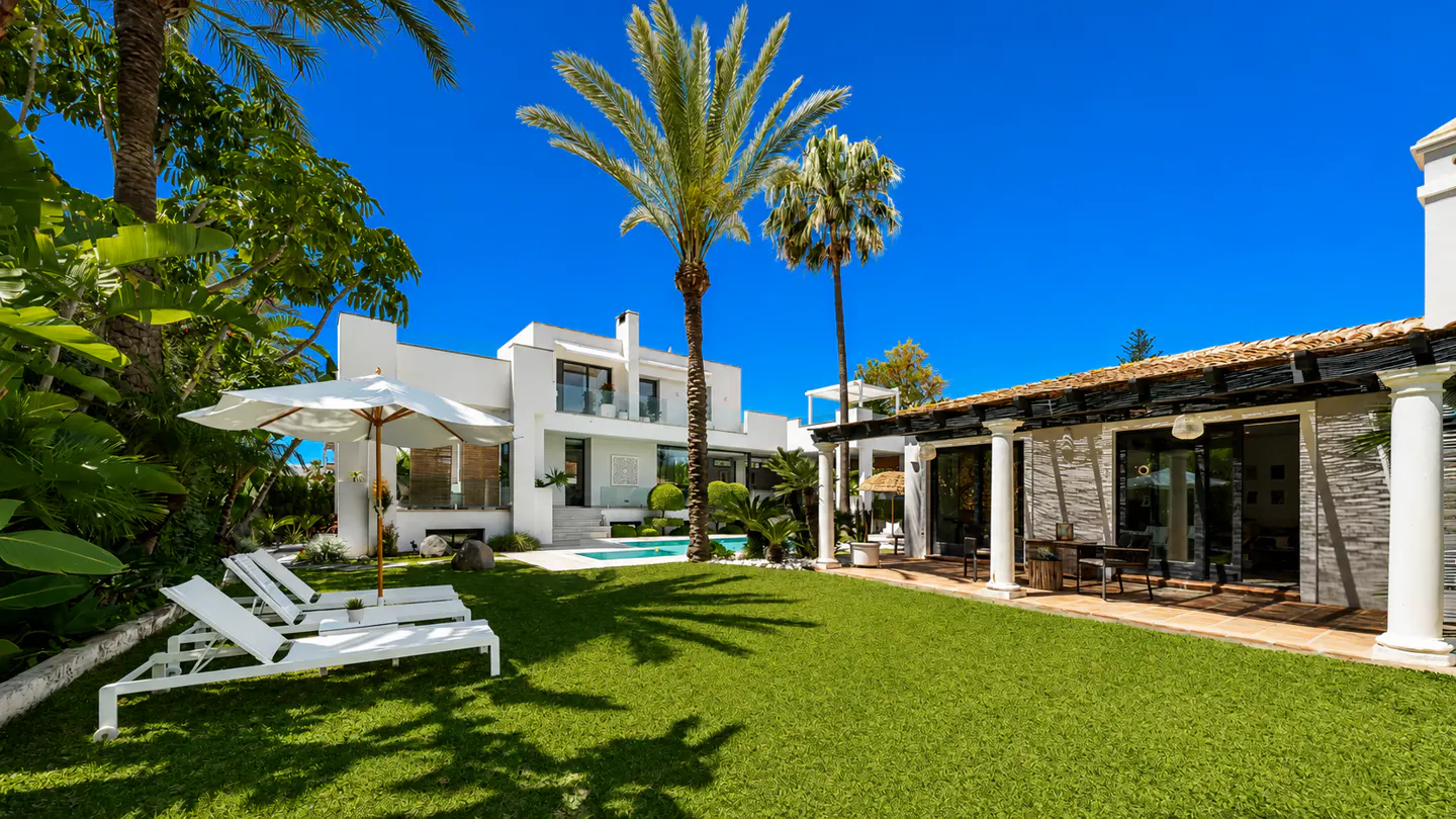 Luxury home exterior with green lawn, palm trees, and blue sky. White lounge chairs and umbrella near a modern white house with a pool.