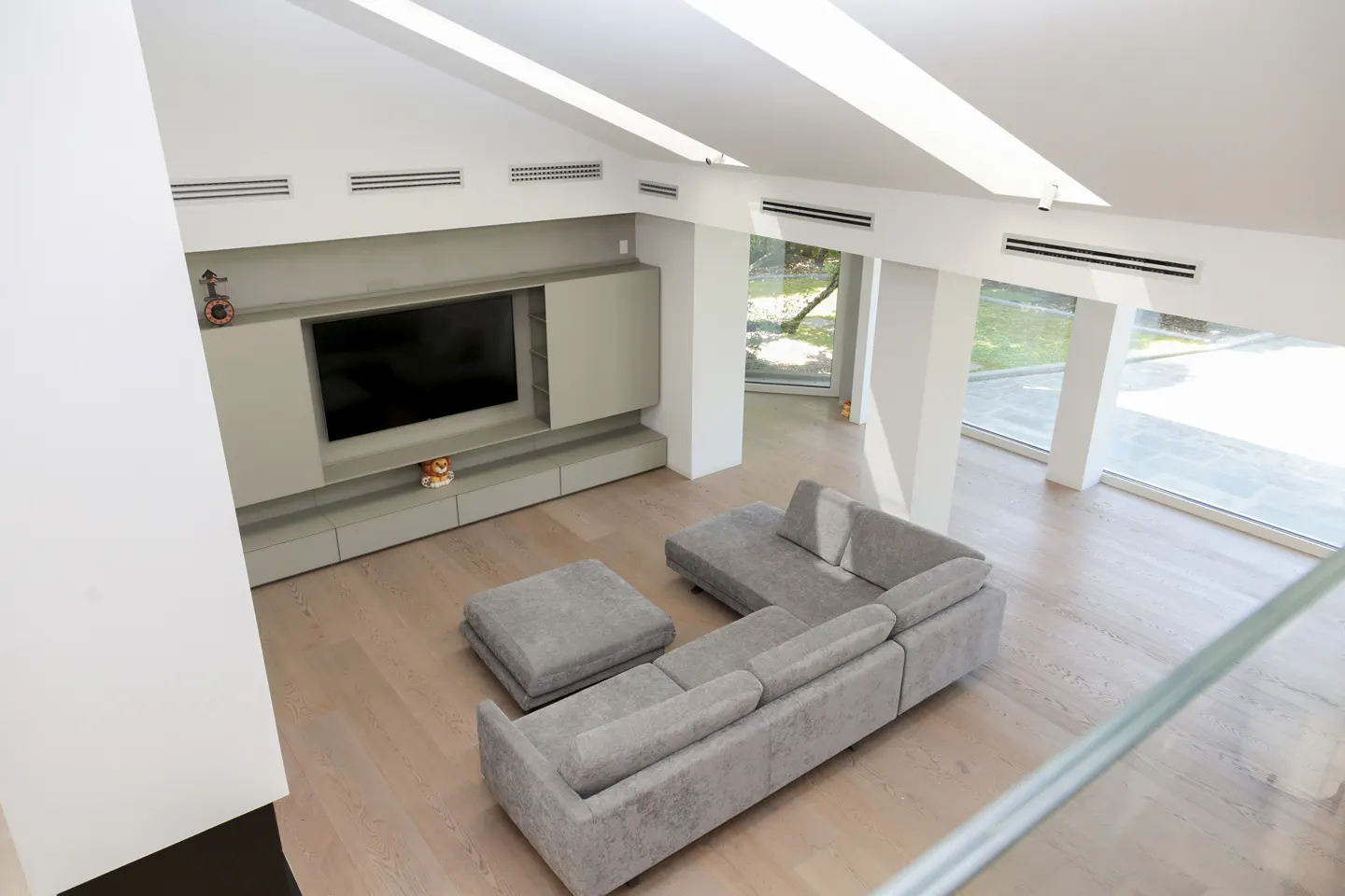 Bright, modern living room with a gray sectional sofa, ottoman, and a large TV on a gray wall unit. Large windows offer a view of greenery.