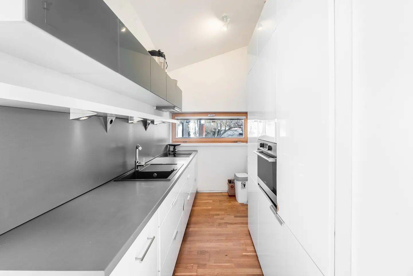 A modern, narrow kitchen with white cabinets, gray countertops, and wood floors. A window is visible at the end of the kitchen.